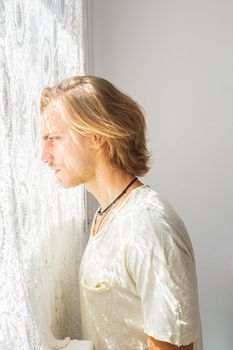 Young man with medium long blonde hair frustrated at not being able to leave home. Loneliness and silence concept. Sun rays coming into the house through the window.