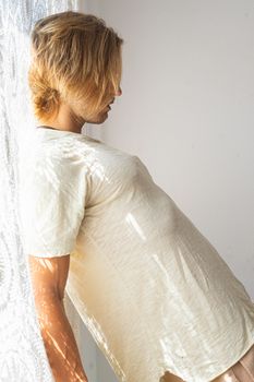 Young man with medium long blonde hair frustrated at not being able to leave home. Loneliness and silence concept. Sun rays coming into the house through the window.