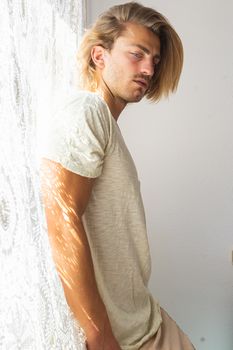 Young man with medium long blonde hair frustrated at not being able to leave home. Loneliness and silence concept. Sun rays coming into the house through the window.