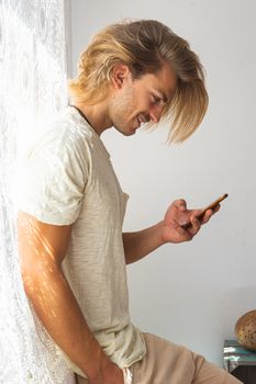 Young man with medium long blonde hair talking on the phone. Sun rays coming into the house through the window. Social distancing and technologies