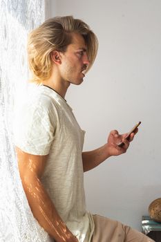 Young man with medium long blonde hair talking on the phone. Sun rays coming into the house through the window. Social distancing and technologies