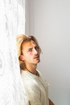 Young man with medium long blonde hair frustrated at not being able to leave home. Loneliness and silence concept. Sun rays coming into the house through the window.