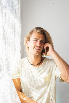 Young man with medium long blonde hair talking on the phone. Sun rays coming into the house through the window. Social distancing and technologies