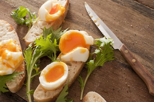 Tasty soft boiled eggs and salad greens on sliced fresh white bread with a kitchen knife on a wooden table