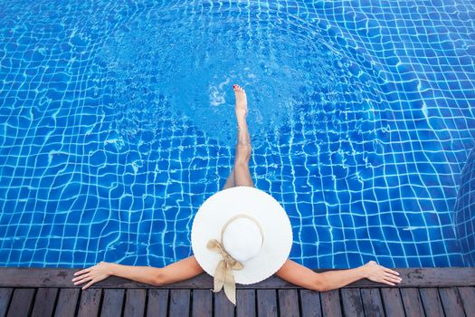 Beautiful woman sunbathing by the pool top view, summer vacation resort background. Girl sitting in swimming pool in big sunhat