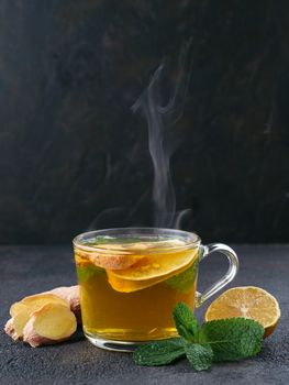 Herbal tea with ginger, mint and lemon. Hot tea glass cup with steam and ingredients on black cement background. Copy space