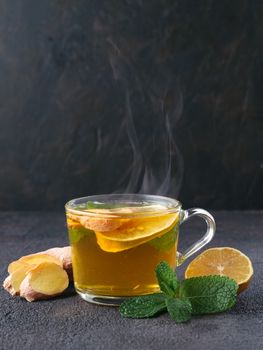 Herbal tea with ginger, mint and lemon. Hot tea glass cup with steam and ingredients on black cement background. Copy space