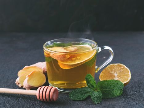 Herbal tea with ginger, mint, lemon and honey. Hot tea glass cup with steam and ingredients on black cement background. Copy space