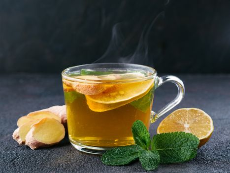 Herbal tea with ginger, mint and lemon. Hot tea glass cup with steam and ingredients on black cement background. Copy space