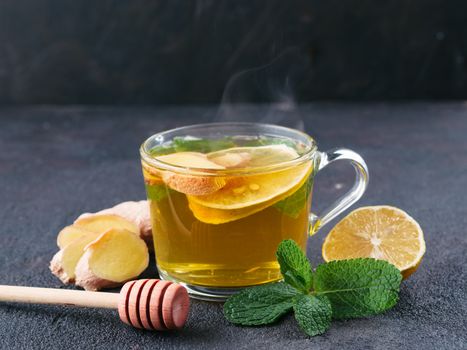 Herbal tea with ginger, mint, lemon and honey. Hot tea glass cup with steam and ingredients on black cement background. Copy space