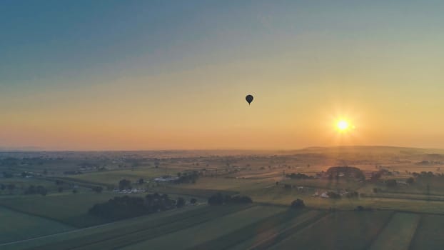 Aerial view of a summer sunset over farm lands with blue skies and red sun and a hot air balloon floating by