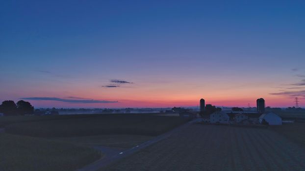 Sunrise over barns, silos and green farmlands with blue, red and orange skies and a few clouds with a mist on the ground