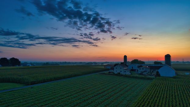 Sunrise over barns, silos and green farmlands with blue, red and orange skies and a few clouds with a mist on the ground