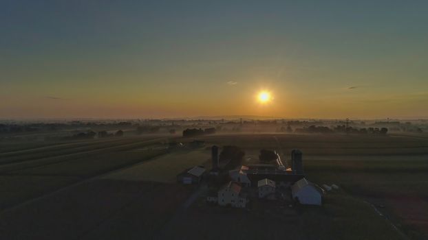 Sunrise over barns, silos and green farmlands with blue, red and orange skies and a few clouds with a mist on the ground