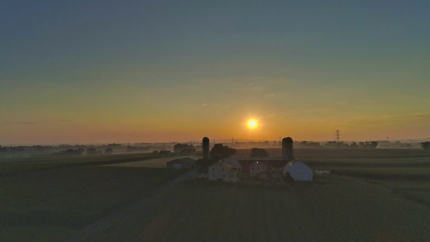 Sunrise over barns, silos and green farmlands with blue, red and orange skies and a few clouds with a mist on the ground