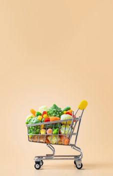 Frozen vegetables assorted in toy shopping cart on cream background. Full of assorted frozen vegetables food shop trolley at beige or yellow backdrop. Minimalistic concept. Vertical. Copy space