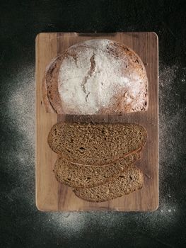 Sliced homemade sourdough rye bread on cutting board over black textured background with rye flour. Top view or flat-lay. Low key