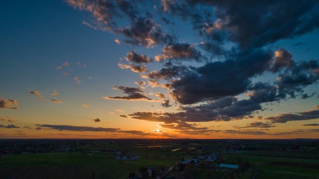 Aerial view of a Sunset over barns, silos and farmlands during the golden hour with blue sky, clouds and red sun