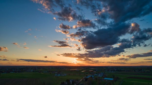 Aerial view of a Sunset over barns, silos and farmlands during the golden hour with blue sky, clouds and red sun