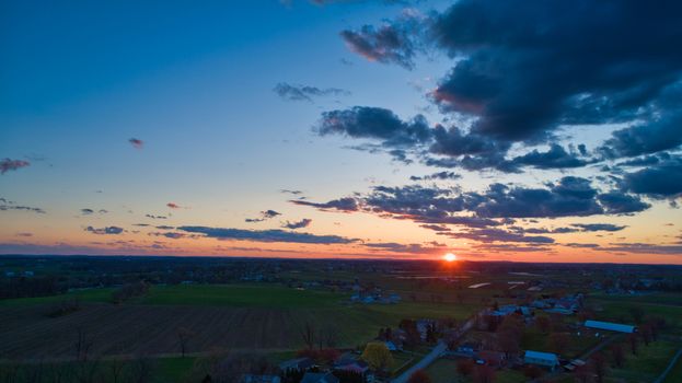 Aerial view of a Sunset over barns, silos and farmlands during the golden hour with blue sky, clouds and red sun