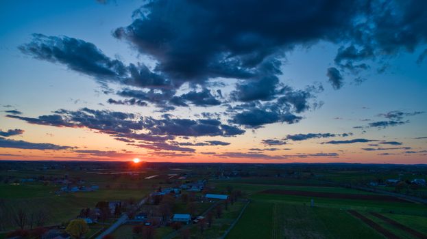 Aerial view of a Sunset over barns, silos and farmlands during the golden hour with blue sky, clouds and red sun