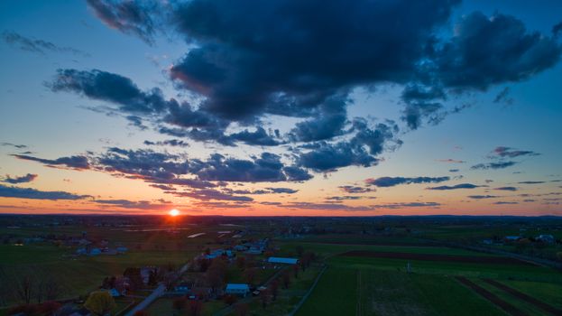 Aerial view of a Sunset over barns, silos and farmlands during the golden hour with blue sky, clouds and red sun