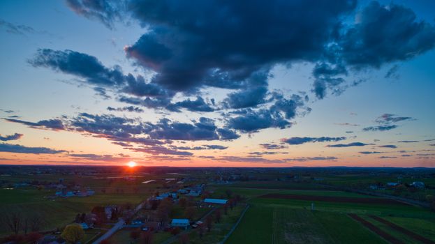 Aerial view of a Sunset over barns, silos and farmlands during the golden hour with blue sky, clouds and red sun