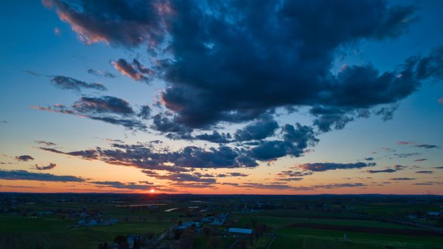 Aerial view of a Sunset over barns, silos and farmlands during the golden hour with blue sky, clouds and red sun
