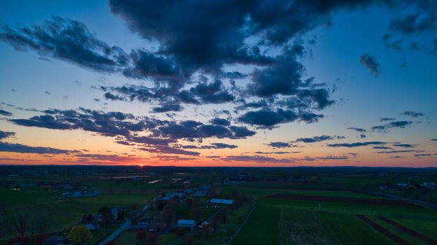 Aerial view of a Sunset over barns, silos and farmlands during the golden hour with blue sky, clouds and red sun