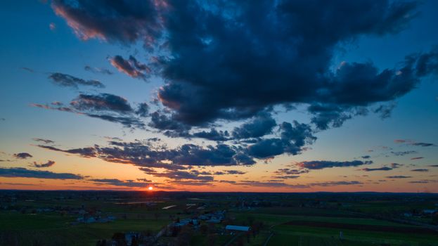 Aerial view of a Sunset over barns, silos and farmlands during the golden hour with blue sky, clouds and red sun