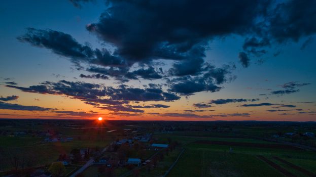 Aerial view of a Sunset over barns, silos and farmlands during the golden hour with blue sky, clouds and red sun