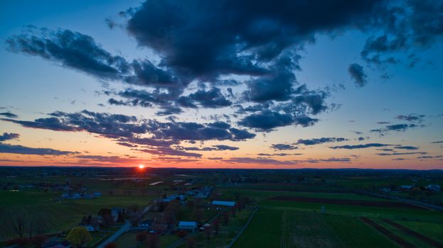 Aerial view of a Sunset over barns, silos and farmlands during the golden hour with blue sky, clouds and red sun