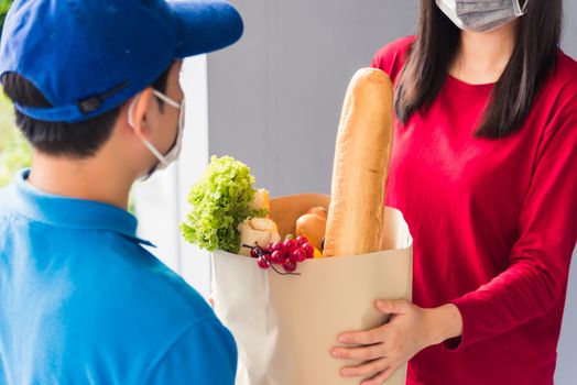 Asian young delivery man in uniform wear protective face mask he making grocery service giving fresh food to woman customer receiving front house under pandemic coronavirus, Back to new normal concept