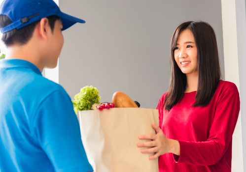 Asian young delivery man in uniform making grocery fast service giving fresh food in paper bag to woman customer receiving at house door under pandemic coronavirus, Back to new normal concept