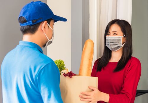 Asian young delivery man in uniform wear protective face mask he making grocery service giving fresh food to woman customer receiving front house under pandemic coronavirus, Back to new normal concept