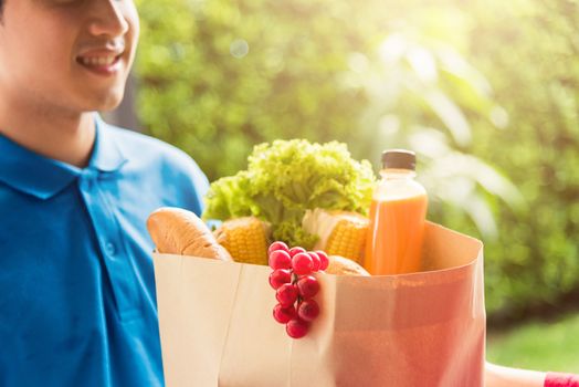 Asian young delivery man in uniform making grocery fast service giving fresh food in paper bag to woman customer receiving at house door under pandemic coronavirus, Back to new normal concept