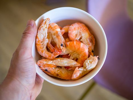 Bowl filled with fresh shrimps in man hands. Chairs are visible in the blurred background. Food for diet - Grilled shrimps with mango salad. Home cook in Asian kitchen.