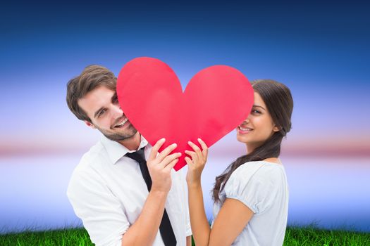 Couple smiling at camera holding a heart against green grass under blue and purple sky