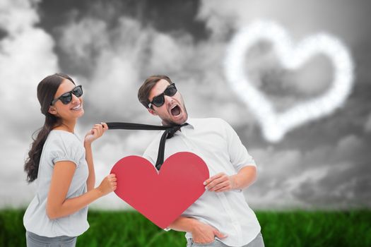 Brunette pulling her boyfriend by the tie holding heart against green grass under grey sky