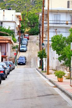 A narrow typical old street of Loutraki in Greece with a cracked road amid parked cars and green trees in the early morning.