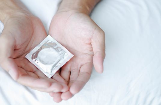 Close up man hand holding condom lying on white bed, health care and medical concept, selective focus