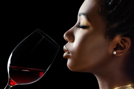 Macro close up low key portrait of sensual african woman smelling red wine.Side view of girl with red wine glass next to face against black background.