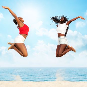 Full  length action portrait of two african girlfriends jumping together on beach. Girls screaming with hands in air.