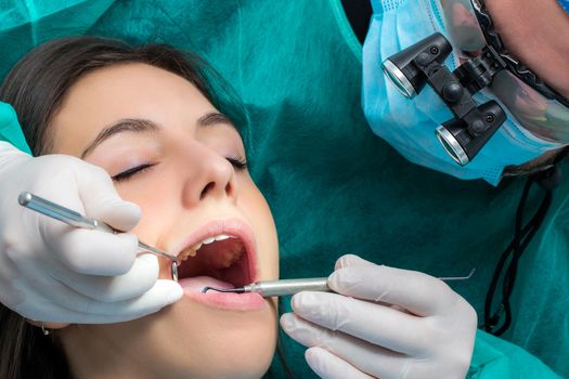 Close up of dentist wearing medical magnifying glasses working on female patient.Surgeon in green gown with mouth mirror and scaler. 