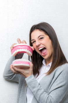 Close up fun portrait of attractive young girl holding oversize human teeth prosthesis.Woman with open mouth against light textured background.