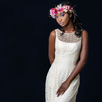 Close up studio portrait of attractive young african bride wearing white wedding gown. Medium shot of girl with colorful flower garland against dark background.