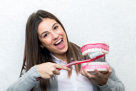 Close up fun portrait of attractive young girl showing oversized human teeth prosthesis.Woman cleaning teeth on prosthesis with toothbrush against light textured background.