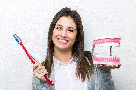 Close up fun portrait of attractive young girl holding oversize toothbrush and human teeth prosthesis.Cute woman standing against light textured background.
