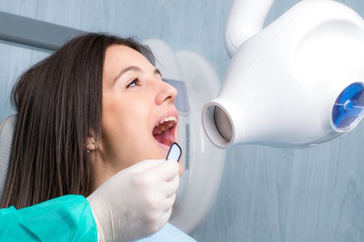 Close up portrait of young woman with open mouth next to x-ray machine in dental clinic.Assistant holding blank x-ray in front of girl.