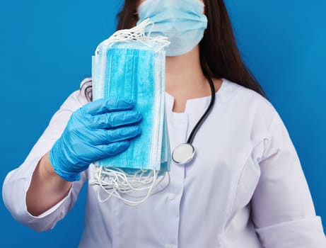 doctor in a white coat and mask holds a stack of protective disposable face masks, physician stands on a blue background